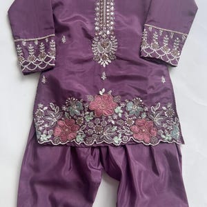 Puede incluir: Un conjunto de t&uacute;nica de manga larga y pantal&oacute;n de color morado con intrincados abalorios y lentejuelas plateadas alrededor del cuello y los pu&ntilde;os. El dobladillo de la t&uacute;nica presenta motivos florales bordados en rosa y azul claro.