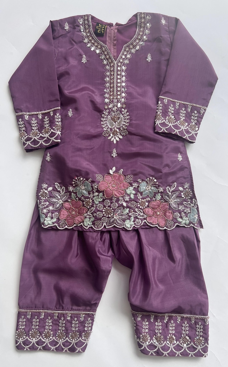 Puede incluir: Un conjunto de t&uacute;nica de manga larga y pantal&oacute;n de color morado con intrincados abalorios y lentejuelas plateadas alrededor del cuello y los pu&ntilde;os. El dobladillo de la t&uacute;nica presenta motivos florales bordados en rosa y azul claro.
