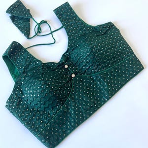 May include: A dark green, sequined top with a matching tie-up accessory. The top features a unique design with a wrap-style front, embellished with small, silver sequins. The neckline and edges are trimmed with a darker green fabric, and it has two decorative buttons.