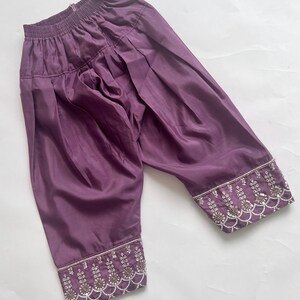Puede incluir: Pantalones de color morado intenso con cintura el&aacute;stica y una silueta fruncida y voluminosa. Los pu&ntilde;os de las piernas est&aacute;n adornados con intrincados bordados plateados, peque&ntilde;as cuentas blancas y lentejuelas en un patr&oacute;n festoneado.