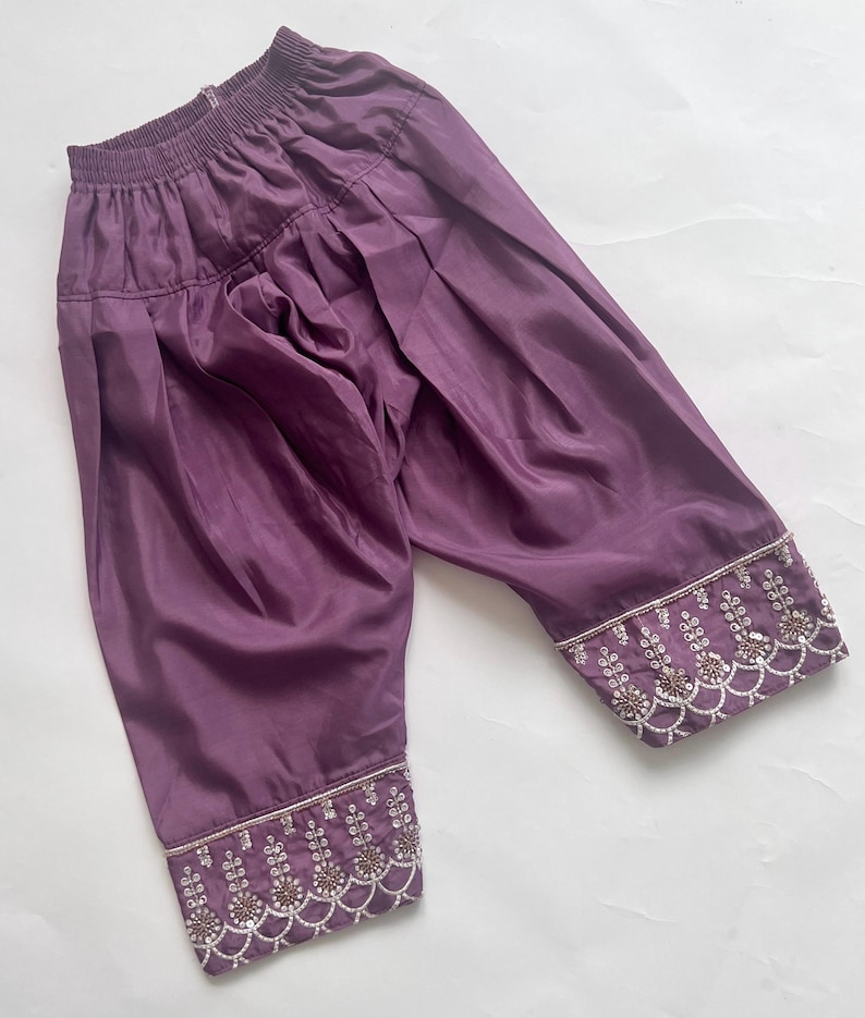 Puede incluir: Pantalones de color morado intenso con cintura el&aacute;stica y una silueta fruncida y voluminosa. Los pu&ntilde;os de las piernas est&aacute;n adornados con intrincados bordados plateados, peque&ntilde;as cuentas blancas y lentejuelas en un patr&oacute;n festoneado.