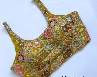 May include: A mustard yellow sleeveless blouse featuring a square neckline and thick shoulder straps. The garment is decorated with intricate multi-coloured embroidery, floral patterns, and metallic sequins. The text Mustard Mehendi is visible.
