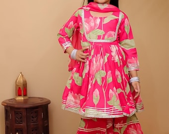 May include: A young girl is wearing a vibrant pink floral print outfit. The ensemble includes a long-sleeved top, matching pants, and a scarf. The outfit is trimmed with silver accents. The girl is smiling, and the background is a neutral beige.