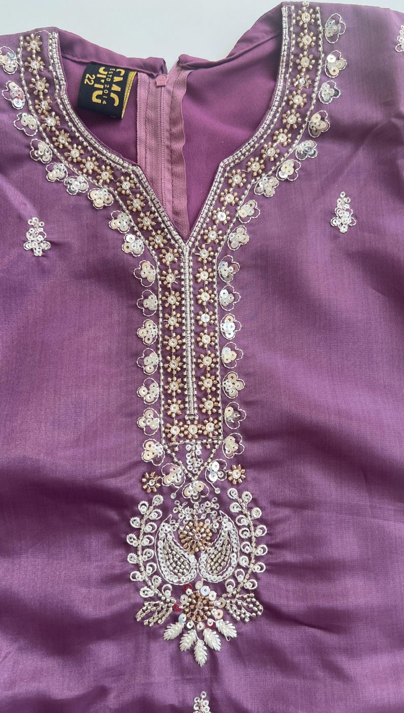 Puede incluir: Una t&uacute;nica de color malva con escote en V adornado con intrincados bordados en plata y oro, lentejuelas transparentes y peque&ntilde;as cuentas. La prenda incluye una etiqueta negra en el cuello con el texto CMC ESTD 2014 22 y una cremallera vertical.