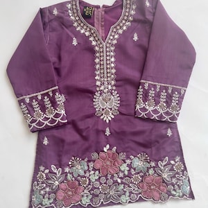 Puede incluir: Una t&uacute;nica morada de manga larga con escote en V. La prenda est&aacute; decorada con lentejuelas plateadas y bordados intrincados alrededor del cuello y los pu&ntilde;os. El dobladillo muestra un patr&oacute;n floral con flores bordadas en rosa y azul claro.