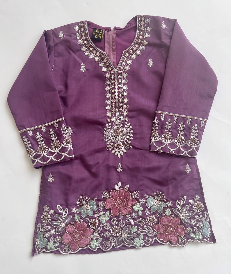Puede incluir: Una t&uacute;nica morada de manga larga con escote en V. La prenda est&aacute; decorada con lentejuelas plateadas y bordados intrincados alrededor del cuello y los pu&ntilde;os. El dobladillo muestra un patr&oacute;n floral con flores bordadas en rosa y azul claro.