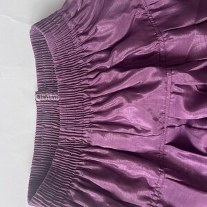 Puede incluir: Una falda de color morado intenso con una cintura el&aacute;stica ancha y fruncida con costuras horizontales visibles. La tela tiene un brillo sutil y una textura suave, con pliegues que caen desde la cintura formando una silueta amplia y fluida.