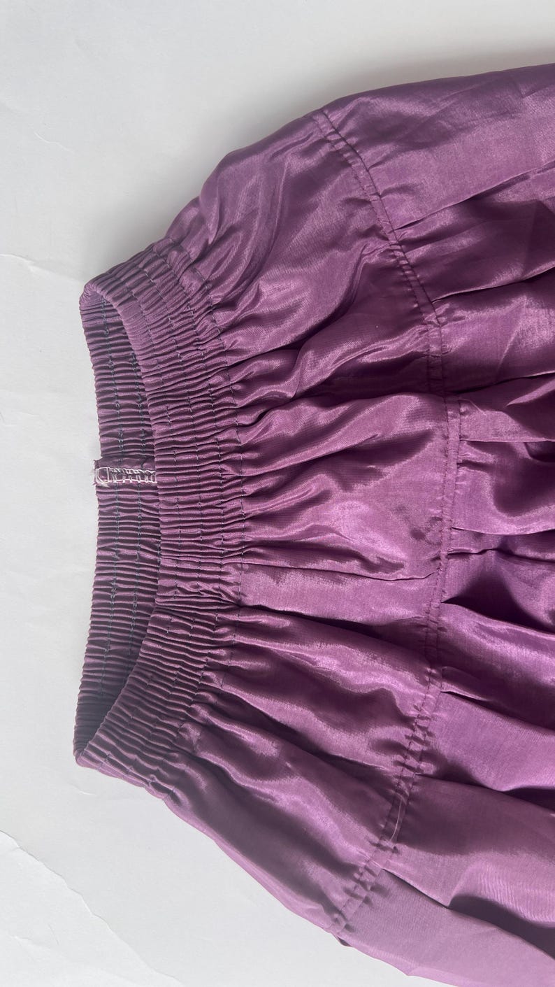 Puede incluir: Una falda de color morado intenso con una cintura el&aacute;stica ancha y fruncida con costuras horizontales visibles. La tela tiene un brillo sutil y una textura suave, con pliegues que caen desde la cintura formando una silueta amplia y fluida.