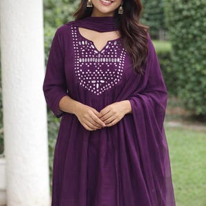 May include: A deep purple tunic and matching scarf set. The tunic features a V-neckline with a decorative mirrored design. The outfit is made of a flowing fabric, and the model is wearing gold earrings.