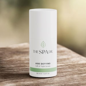 May include: A white cylindrical bottle of The Spa Dr. Age Defying Lift & Tight Serum. The label includes the brand name, product name, and a green band at the bottom with the volume, 1 US fl. oz. The background is a blurred wooden surface.