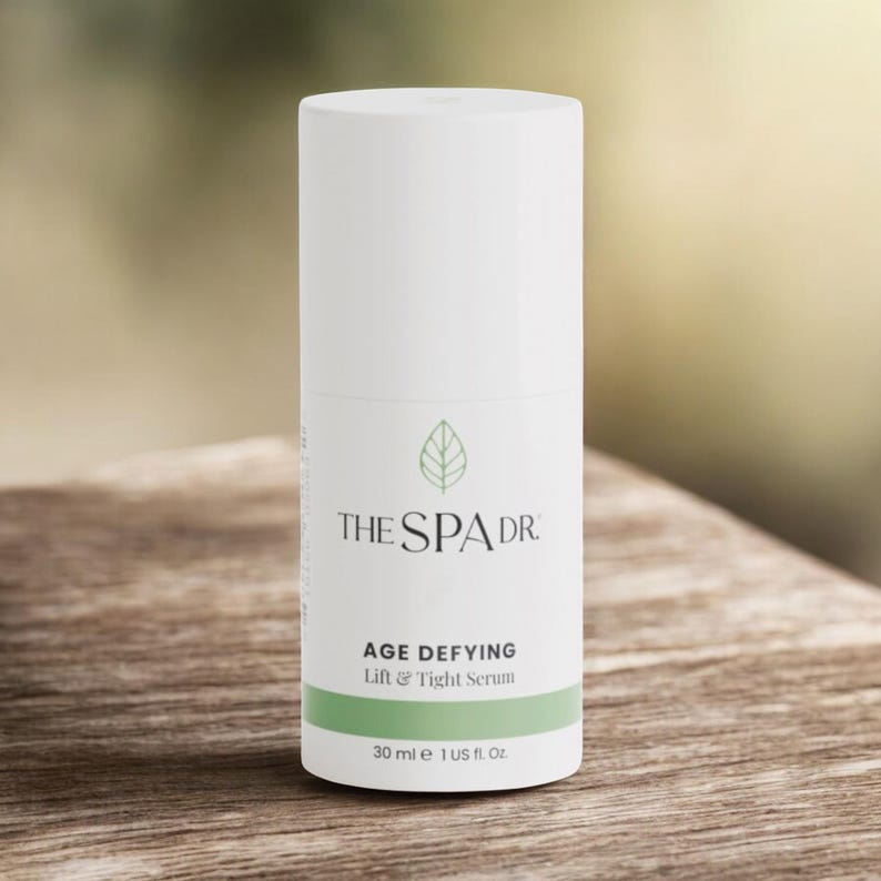 Op de afbeelding: Een witte cilindrische fles The Spa Dr. Age Defying Lift & Tight Serum. Het etiket bevat de merknaam, productnaam en een groene band aan de onderkant met het volume, 30 ml. De achtergrond is een vage houten ondergrond.