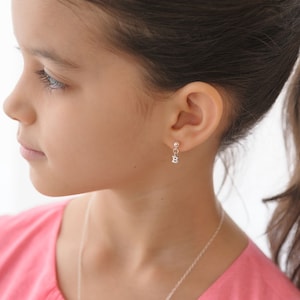 May include: A young person wearing a pink shirt with a silver initial 'B' charm earring.