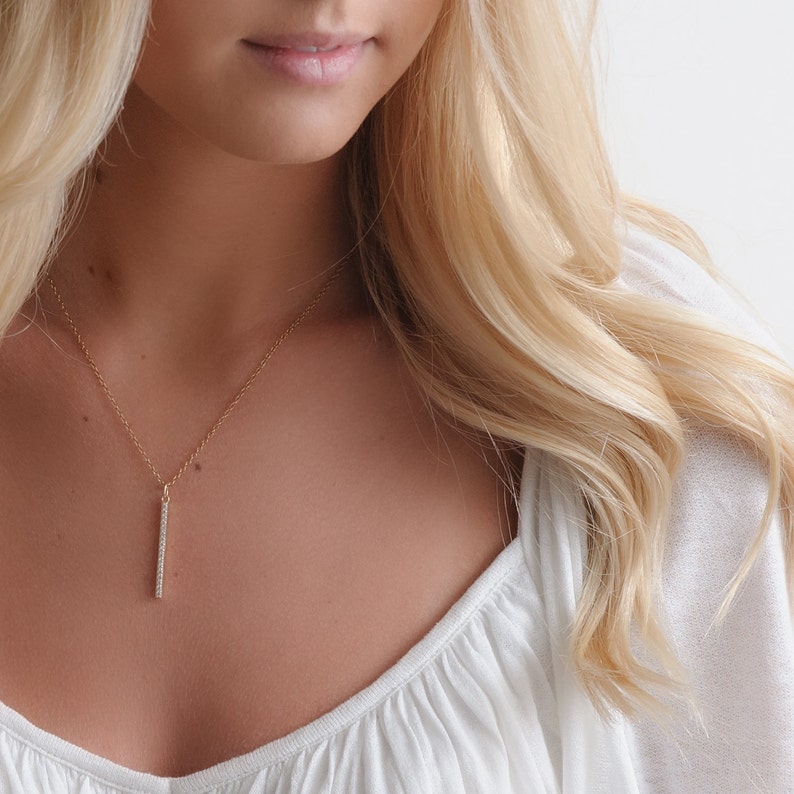 Pu&ograve; includere: Una collana con catena d'oro con un delicato ciondolo a barra verticale incastonato con piccole pietre trasparenti.