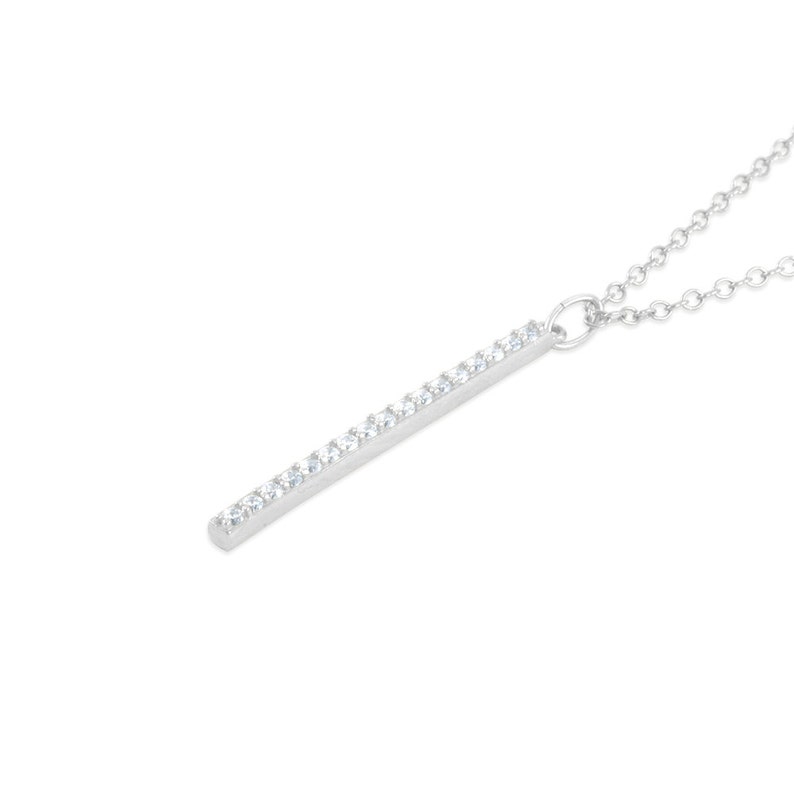 Pu&ograve; includere: Una collana d'argento con un ciondolo lungo e sottile a forma di barra incrostata di piccoli cristalli di zircone cubico trasparenti.