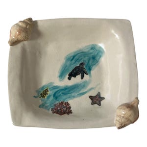Puede incluir: Plato cuadrado de cerámica con base color crema. El plato presenta una escena oceánica pintada a mano con una tortuga marina, una estrella de mar y coral. Dos conchas marinas están adheridas a las esquinas, añadiendo un tema costero. Aproximadamente 20 cm x 20 cm.
