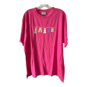 Puede incluir: Una camiseta rosa brillante con cuello redondo y mangas cortas. La camiseta presenta un diseño caprichoso de ropa colgada en un tendedero, que incluye un vestido, pantalones y una camisa. La etiqueta dice "Tide & True".