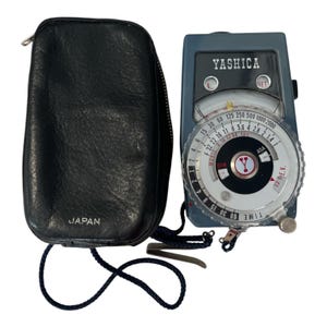 May include: A vintage Yashica light meter with a black leather case. The meter is gray with a circular dial and the brand name "YASHICA" on top. The case is black and has the word "JAPAN" printed on it.