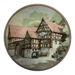 May include: Round decorative plate featuring a detailed watercolor painting of a traditional European village scene. The plate depicts a half-timbered building with a red tile roof, two figures, and a small bench. The plate has a brown border.