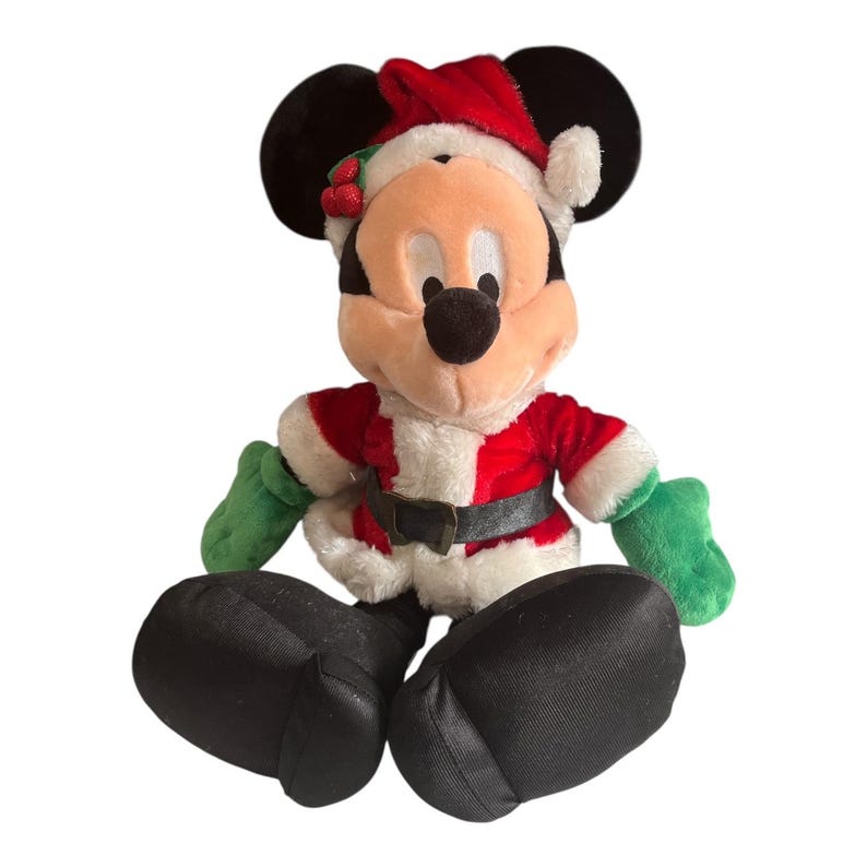 Puede incluir: Un peluche de Mickey Mouse vestido con un traje de Pap&aacute; Noel. La figura lleva un traje rojo y blanco con un cintur&oacute;n negro, guantes verdes y un gorro de Pap&aacute; Noel. El peluche tiene orejas negras y pies grandes negros.