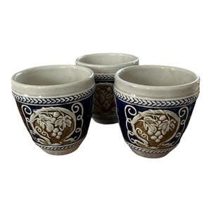 Puede incluir: Tres tazas de cerámica con esmalte azul profundo y un medallón central en relieve de color tostado que representa un racimo de uvas y hojas de vid. Los bordes y bases tienen un patrón trenzado blanco y el interior es de color blanco roto.