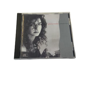 May include: A compact disc in a clear plastic jewel case with a black spine. The cover art features a black and white portrait of Gloria Estefan with the text "gloria estefan cuts both ways" printed in red and black on a gray background.