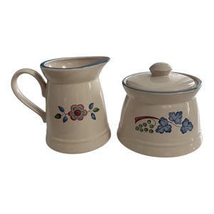 May include: Cream-colored ceramic creamer and sugar bowl set. The creamer has a handle and a floral design. The sugar bowl has a lid and a grape design. Both pieces have blue trim.