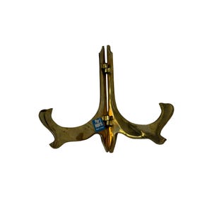 May include: A gold-toned metal display stand with a hinged design and two curved arms for holding items. A small blue rectangular sticker with the white text Pier 1 Imports is attached to the center of the stand, which features a polished finish.