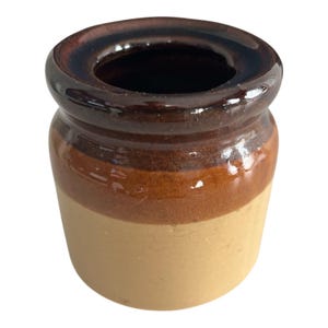 May include: A small, ceramic jar with a dark brown rim and interior. The jar has a light tan base, a medium brown band, and a darker brown band near the top. The jar is likely for storage or decoration.