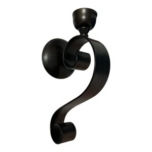 Könnte beinhalten: Eine dunkelbronze Wandleuchte mit einem dekorativen, geschwungenen Design. Die Leuchte hat eine kreisförmige Rückplatte, eine Kerzenschale und ein stilisiertes, scrollartiges Element. Die Metallhalterung ist für eine einzelne Kerze konzipiert.