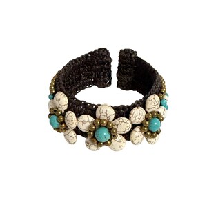 May include: A dark brown crocheted cuff bracelet featuring three flower-shaped embellishments. Each flower consists of white crackle-patterned oval beads as petals surrounding a central round turquoise bead and small metallic bronze-toned beads.