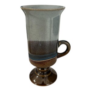 May include: A tall, ceramic mug with a flared rim and a handle. The mug features a color gradient, transitioning from a light blue-gray at the top to a dark brown at the base, with a blue accent. The base is a pedestal shape.