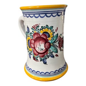 Puede incluir: Taza de cerámica blanca con asa, decorada con coloridos motivos florales. La taza tiene un borde y una base amarillos, con ribete festoneado azul. Las flores son rojas, amarillas y azules, con hojas verdes. Una pieza decorativa para la cocina.