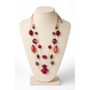 May include: A three-strand necklace displayed on a mannequin. The necklace features various sizes and shapes of red, pink, and purple beads. The beads are strung on thin, gold-colored chains. The necklace is a statement piece.