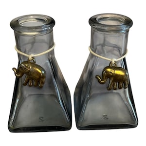 May include: Two gray glass vases with a trapezoidal shape and a round opening. Each vase has a gold-colored elephant charm hanging from a string around the neck. The vases are empty and sit side by side.