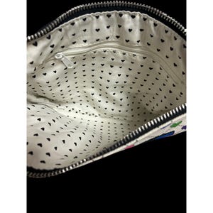 Op de afbeelding: Open zicht op een witte clutch met een zwarte rits en een zwarte hartpatroonvoering. De buitenkant heeft een kleurrijk bloemenontwerp. De tas is open en toont de binnenkant.