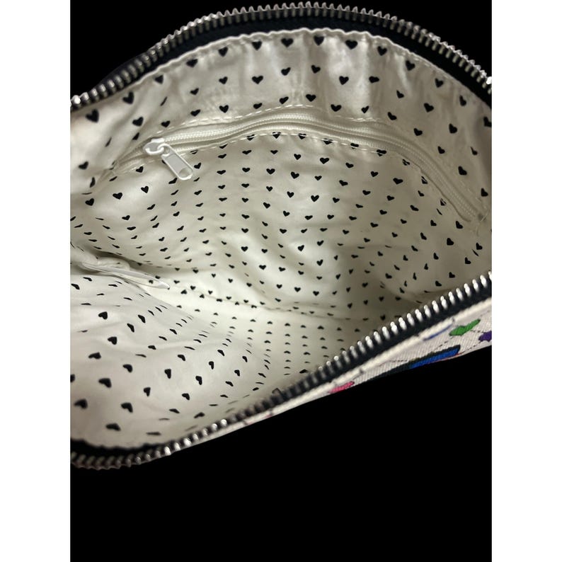 Op de afbeelding: Open zicht op een witte clutch met een zwarte rits en een zwarte hartpatroonvoering. De buitenkant heeft een kleurrijk bloemenontwerp. De tas is open en toont de binnenkant.