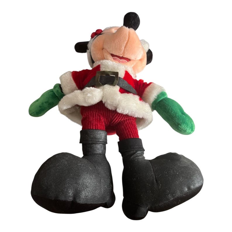 Puede incluir: Un personaje navide&ntilde;o de peluche con un traje de Pap&aacute; Noel rojo con ribetes blancos, guantes verdes y botas negras grandes. El personaje tiene una cara sonriente, orejas negras y un peque&ntilde;o lazo rojo. Una decoraci&oacute;n festiva.