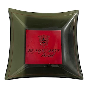 May include: Square, olive-green glass dish with a red center. The dish has a slightly curved shape and a thin red border. The center features the words "Beaux Arts Hotel" in white script.