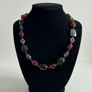 Puede incluir: Un collar de cuentas multicolores exhibido en un maniquí negro. El collar presenta cuentas facetadas en tonos rojos, verdes, morados y grises, unidas con eslabones de metal plateado. Las cuentas varían en forma y tamaño.