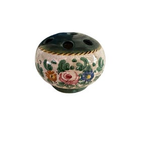 May include: A round ceramic flower frog with a dark green top featuring multiple circular openings. The body has a cream-colored base decorated with hand-painted green leaves and floral motifs in shades of pink, orange, and blue.