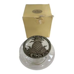 May include: A clear glass jar with a silver-colored pineapple lid, presented with its original yellow box. The lid features a detailed pineapple design within a decorative border. The box has a label.