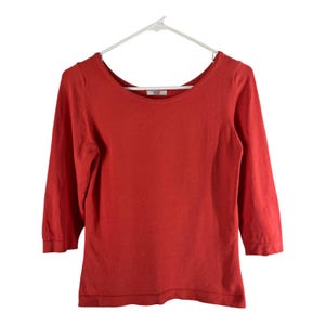 Puede incluir: Un suéter rojo con cuello redondo y mangas tres cuartos cuelga de una percha blanca. El suéter tiene un diseño simple y clásico y está hecho de un material de punto suave. La marca "Mossimo" es visible en la parte posterior del cuello.