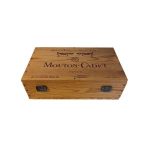May include: A light brown wooden wine box, featuring the words "Mouton Cadet" in dark brown. The box has two black latches and the words "Baron Philippe de Rothschild" on the top. The box is designed to hold two bottles of wine.
