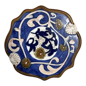 May include: A decorative ceramic plate with a dark brown rim and a blue background. The plate features white and pink floral designs, brown flower accents, and a central white circle with dark blue calligraphy.