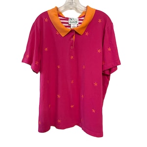 May include: A hot pink polo shirt with an orange collar and embroidered orange star patterns. The collar has a striped pattern on the inside. The shirt has short sleeves and three buttons.