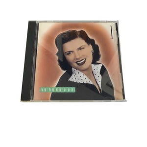 Puede incluir: Una caja de disco compacto con el retrato de una persona de cabello oscuro, vistiendo una prenda negra con cuello de lunares blancos. La portada incluye el texto Honky Tonk Merry Go Round en un recuadro verde sobre un fondo marrón claro.