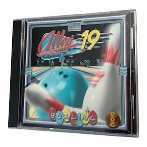 Puede incluir: Una caja de CD con el juego "Alley 19 Bowling". La portada muestra una bola de bolos, bolos y un jugador. El título del juego está en una fuente estilizada con un esquema de color rojo y blanco. La caja del CD está en una caja de plástico transparente.