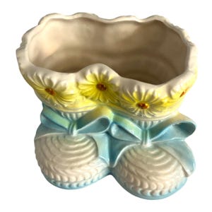 Könnte beinhalten: Keramik-Pflanzgefäß in Form von Babyschuhen. Der Pflanztopf ist weiß mit gelben Gänseblümchen-Akzenten und hellblauen Bändern. Der Innenraum ist cremefarben.