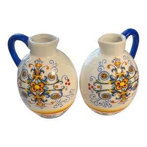 May include: Two white ceramic pitchers with blue handles. Each pitcher features a floral design in orange, yellow, and blue. The pitchers have a rounded shape and a decorative band around the base.