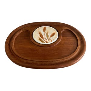 Op de afbeelding: Ovale houten serveerschaal met een verzonken midden en een decoratief keramisch inzetstuk. Het inzetstuk heeft een tarwearen-ontwerp in bruin-, goud- en crèmetinten. De schaal heeft een warme bruine kleur.